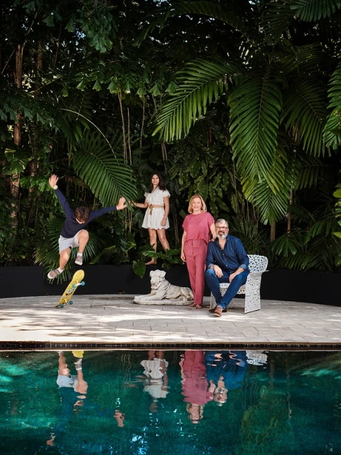Group portrait near a swimming pool, featuring a skateboarder mid-air, two standing women, and a seated man, set against a lush green backdrop.