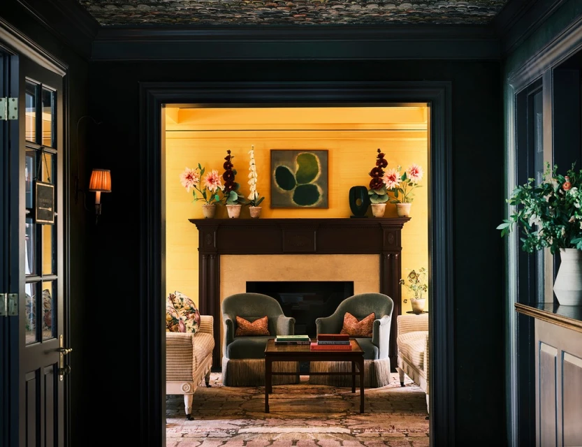 Interior view through a dark doorway reveals a sunlit room with a fireplace, green upholstered armchairs, floral arrangements, and art.