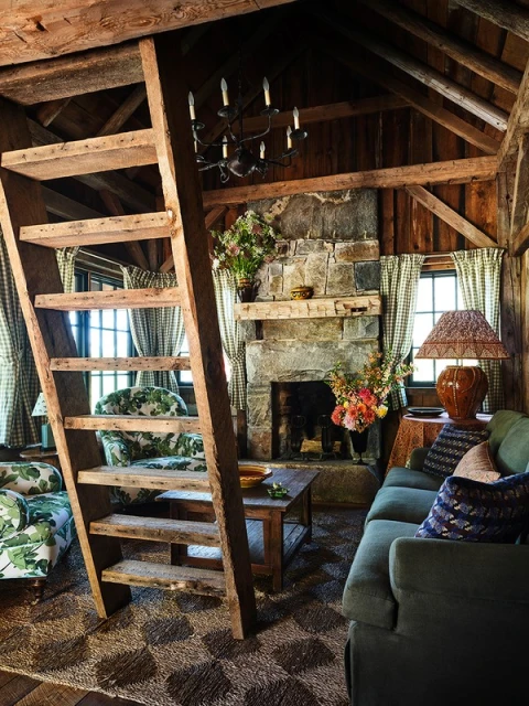 Cozy rustic interior with stone fireplace, exposed beams, wooden ladder, patterned armchairs, and a green sofa creating a warm atmosphere.