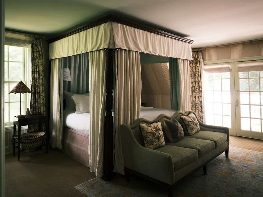 A four-poster bed with white curtains and a light green sofa with pillows in a well-lit bedroom with soft lighting and a carpet.