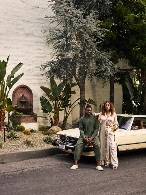 Outdoor portrait of a man in green and woman in cream-colored outfit posing in front of vintage convertible with lush greenery.