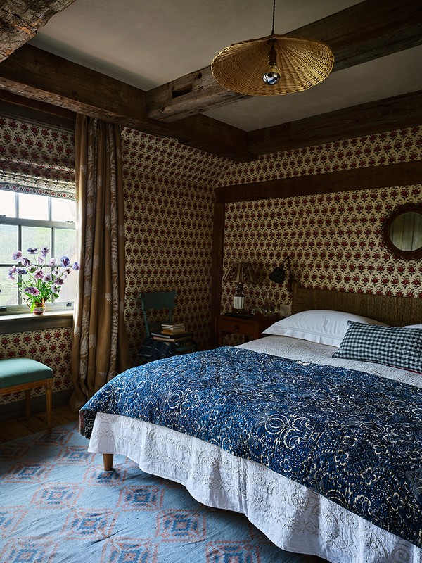 Bedroom interior with a bed featuring a blue patterned quilt, patterned wallpaper, natural light from a window, and wooden ceiling beams.