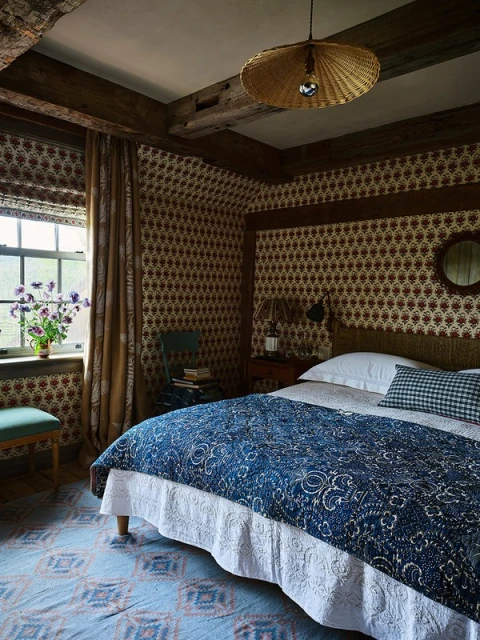 Bedroom interior with a bed featuring a blue patterned quilt, patterned wallpaper, natural light from a window, and wooden ceiling beams.