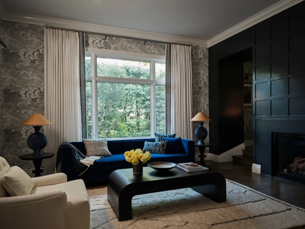 Interior living room with blue velvet sofa, dark wood coffee table, white armchairs, and large multi-pane window with lush greenery view.