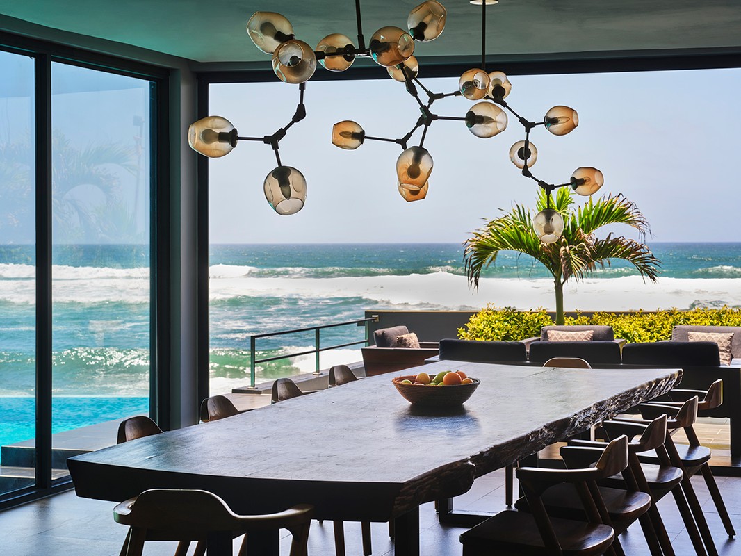 Interior dining area with a long wood table and chairs, modern lighting, ocean view through large windows, and an outdoor lounge space.
