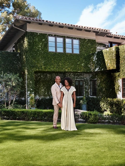 A couple poses in front of a ivy-covered house on a sunny day. The woman wears a dress, while the man wears a tan suit.