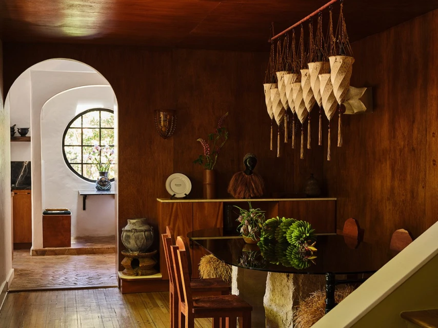 Interior shot of a dining room featuring a round table, wooden chairs, and a distinctive chandelier against wood-paneled walls.