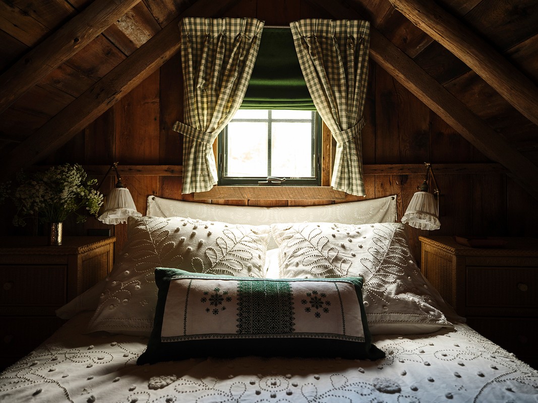 Cozy bedroom interior with a bed covered in embroidered white linens, patterned curtains, warm wood walls, and soft, diffused lighting.