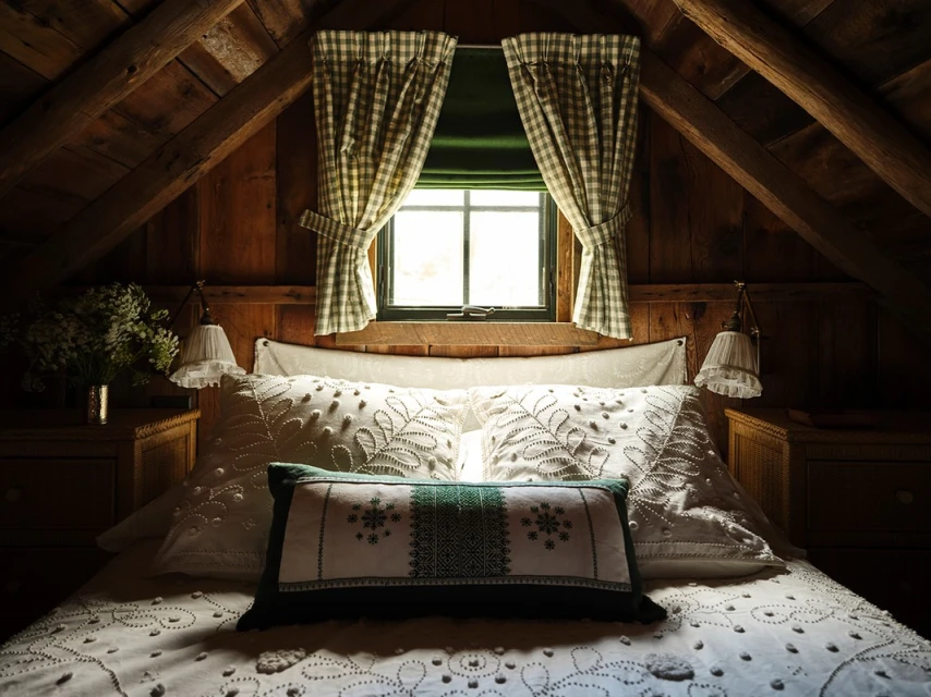 Cozy bedroom interior with a bed covered in embroidered white linens, patterned curtains, warm wood walls, and soft, diffused lighting.