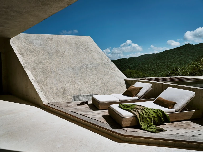 Outdoor terrace featuring two lounge chairs with cushions and throw pillows, set on a wooden deck with a mountain backdrop and blue sky.