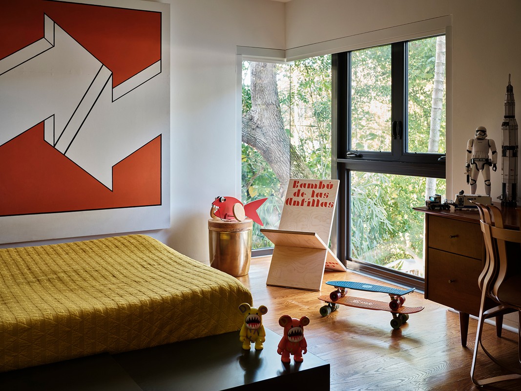 A child's bedroom with an art piece, bed, two skateboards, and toys, with a view of a forest through a large window.