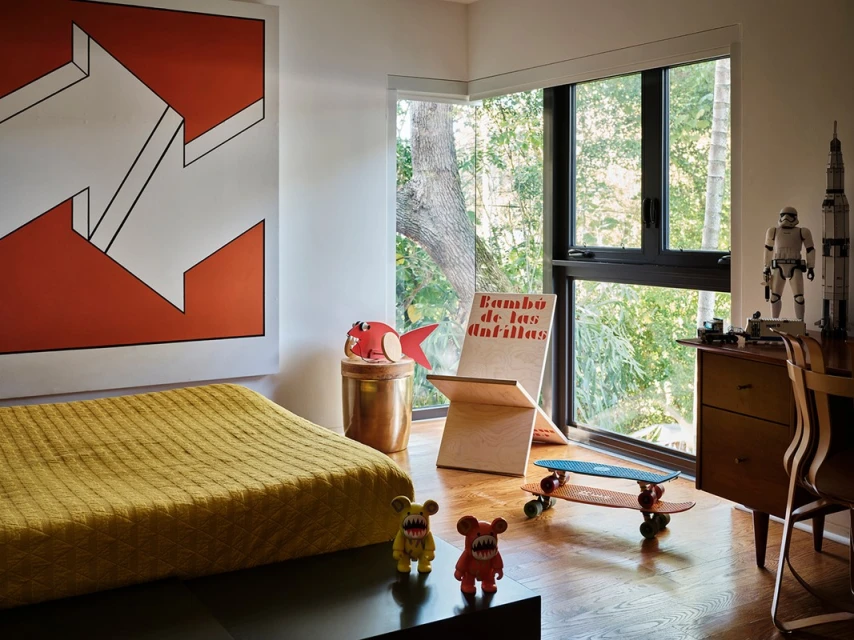 A child's bedroom with an art piece, bed, two skateboards, and toys, with a view of a forest through a large window.
