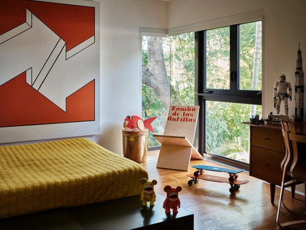 A child's bedroom with an art piece, bed, two skateboards, and toys, with a view of a forest through a large window.