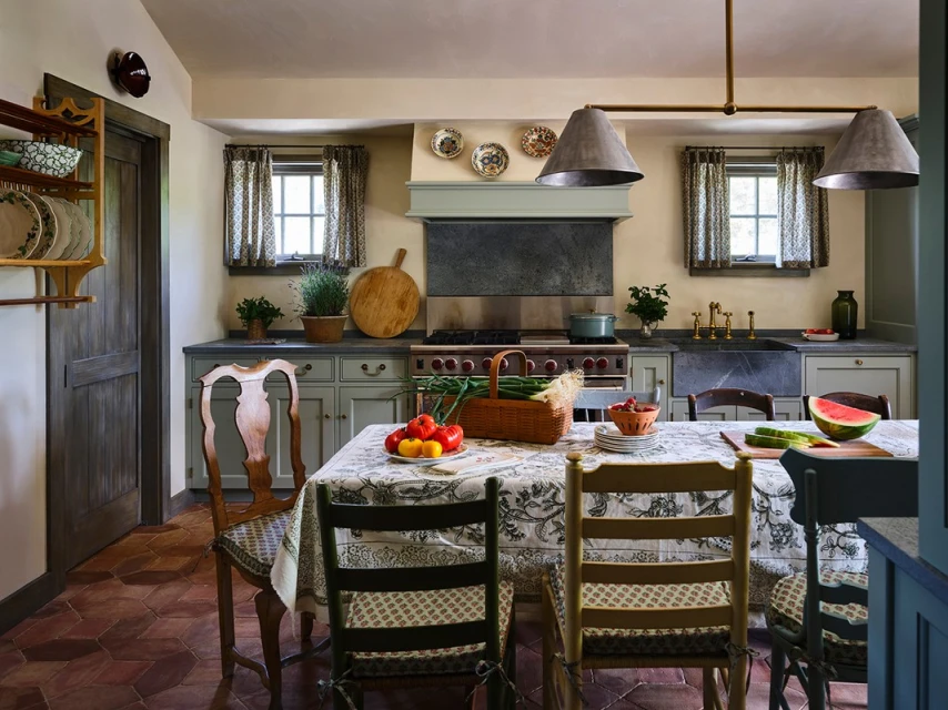 Country kitchen interior with a rustic table centerpiece, light green cabinets, terracotta tile floor, and hanging metal pendant lights.