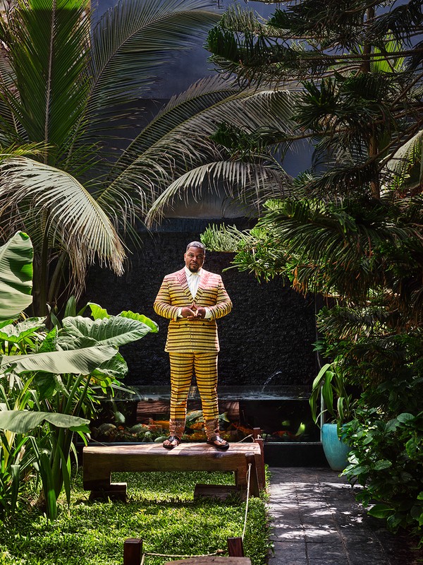 Adult male in a yellow-striped suit stands in a lush garden setting, surrounded by tropical plants and trees, posing on a rustic wooden bench.
