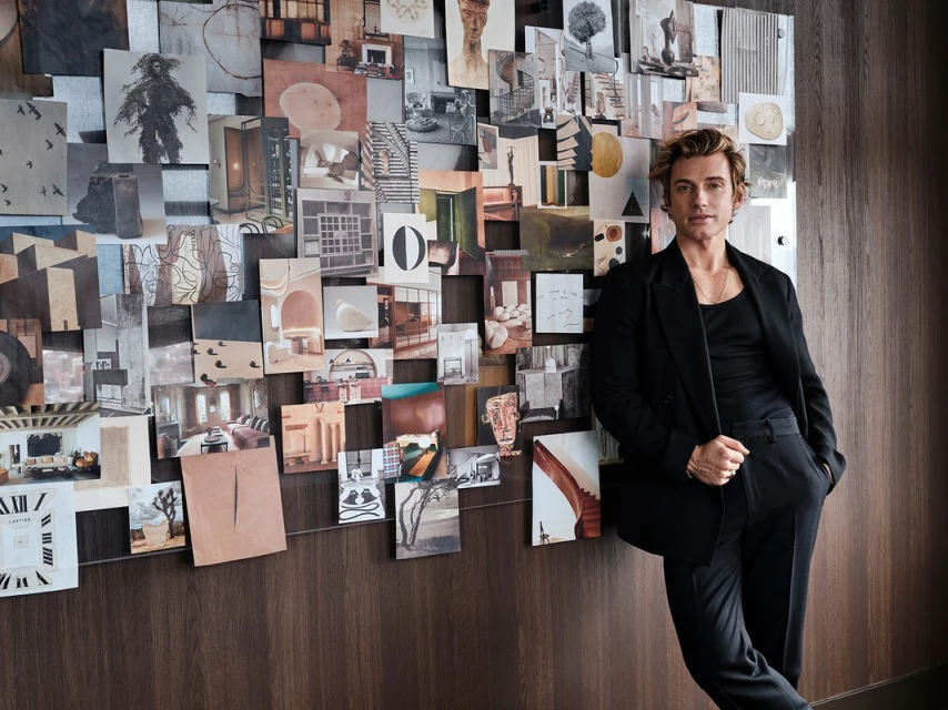 Adult male in black suit leans against a wall covered with a collage of photographs in an indoor setting with soft lighting.