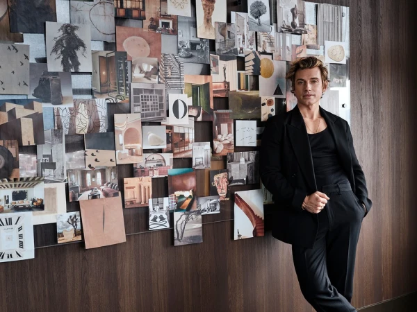 Adult male in black suit leans against a wall covered with a collage of photographs in an indoor setting with soft lighting.