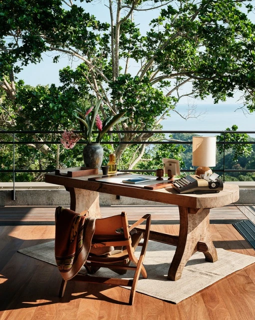Rustic wooden desk setup on a balcony with trees and ocean in the background, featuring a vintage typewriter and modern decor.