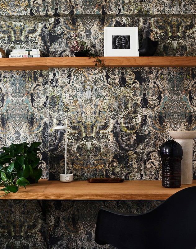 Interior home office with a wooden desk, black chair, modern lamp, decor objects, and patterned wallpaper in a well-lit indoor space.