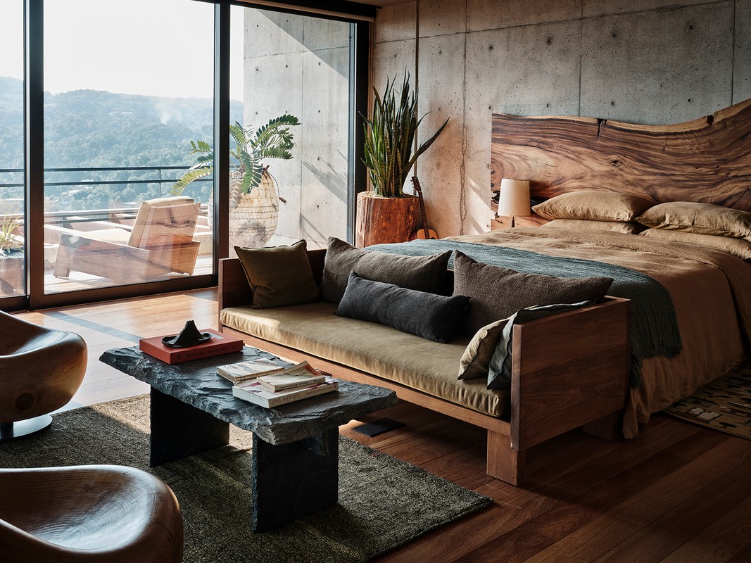 Modern bedroom interior with a wooden bed, concrete walls, stone coffee table, and a view to a lush green, mountainous landscape.