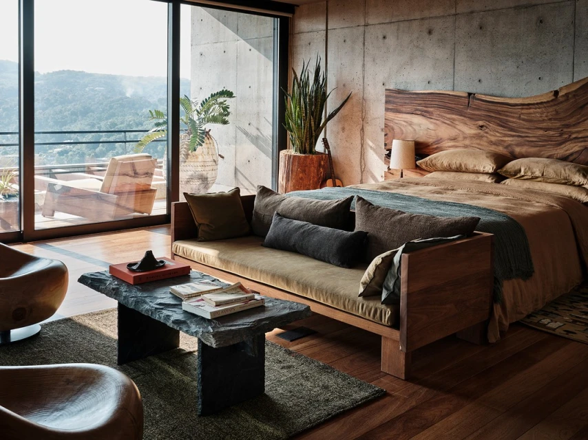 Modern bedroom interior with a wooden bed, concrete walls, stone coffee table, and a view to a lush green, mountainous landscape.