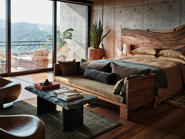 Modern bedroom interior with a wooden bed, concrete walls, stone coffee table, and a view to a lush green, mountainous landscape.