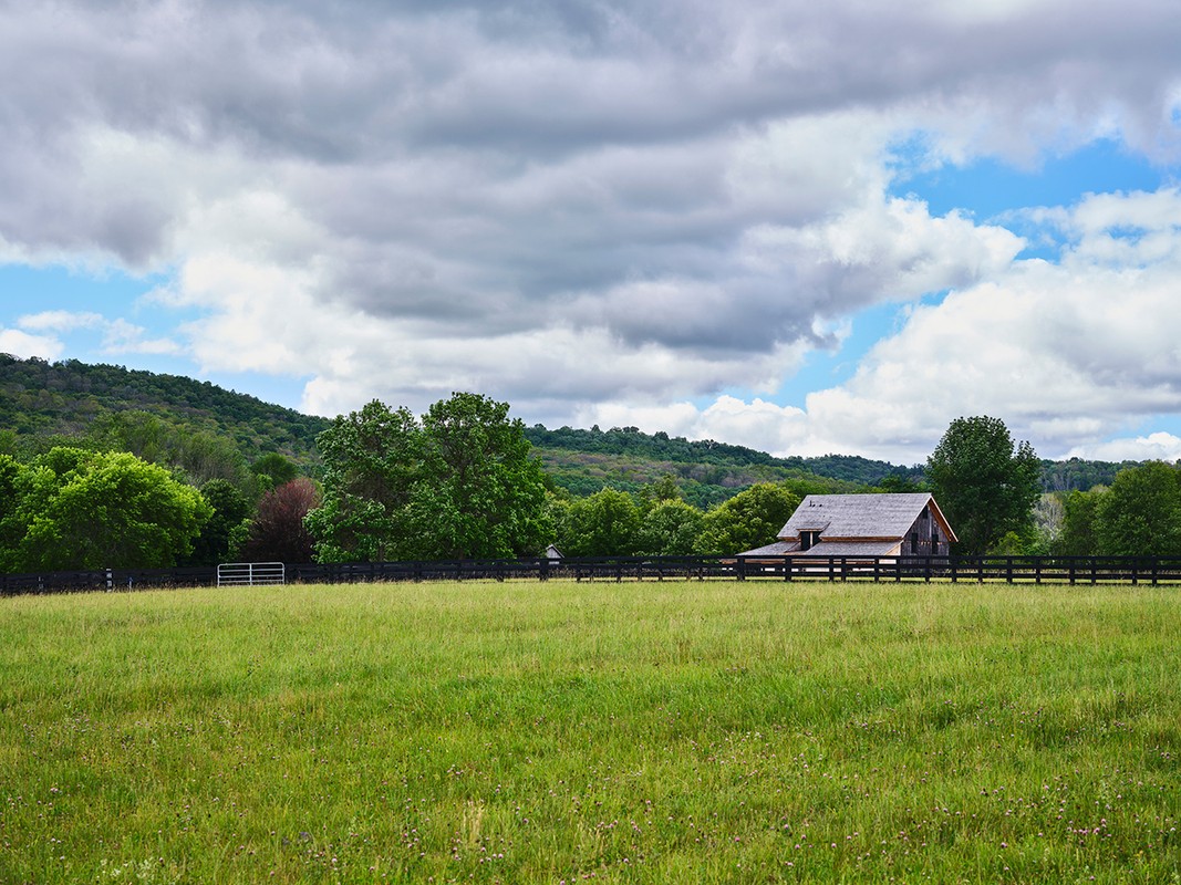 Open green field with a black fence and barn with trees and a hill in the background under a sky with many clouds.