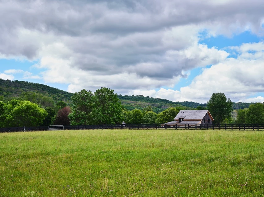 Open green field with a black fence and barn with trees and a hill in the background under a sky with many clouds.