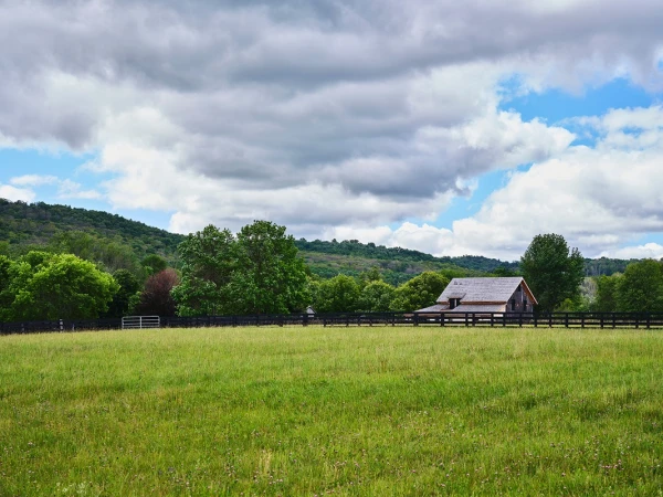 Open green field with a black fence and barn with trees and a hill in the background under a sky with many clouds.