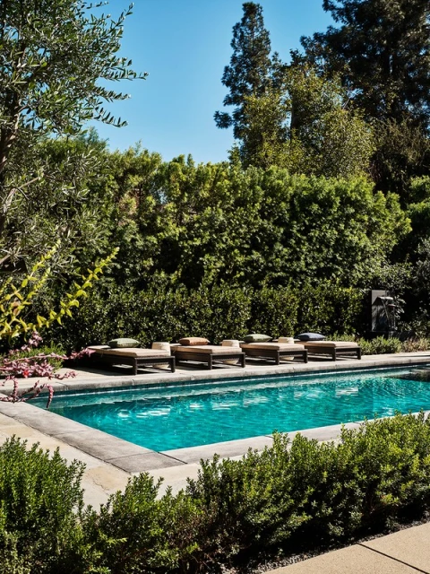 An outdoor swimming pool surrounded by lush green foliage with lounge chairs lined up along the pool's edge, beneath a clear blue sky.