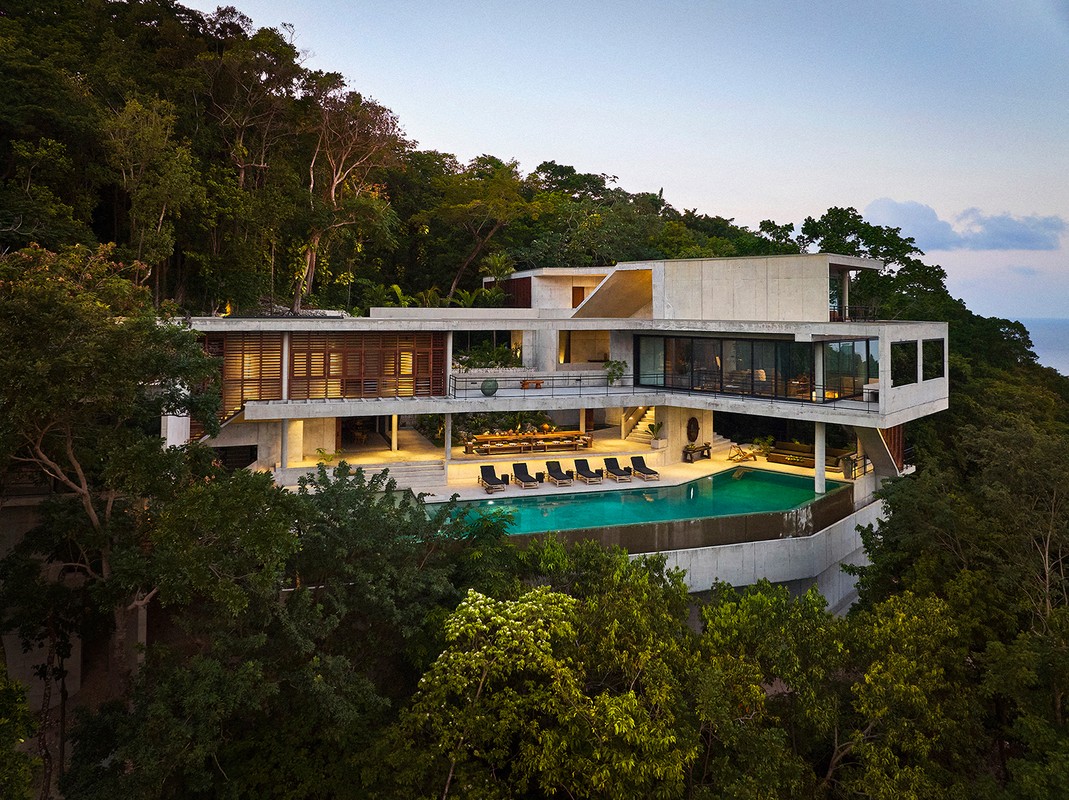 Exterior view of a modern concrete house featuring a pool and outdoor lounge, set within a lush, tropical hillside landscape.