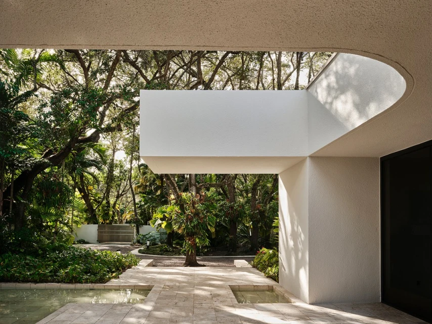 Outdoor view of modern building exterior with white concrete walls, water feature, and lush trees under natural light. Architecture design.
