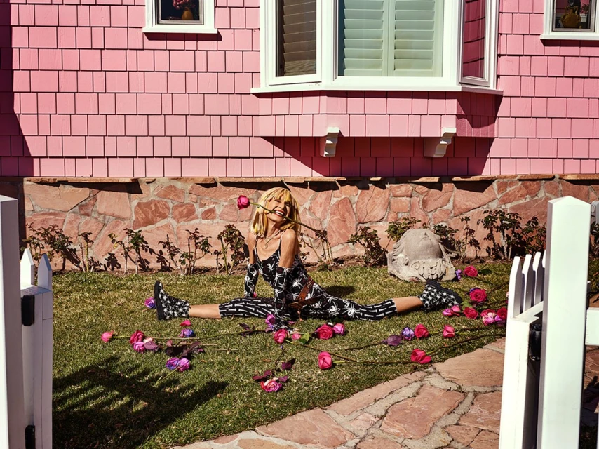 A young woman in a patterned catsuit performs a split on the lawn of a pink house surrounded by fallen roses in the bright sunlight.