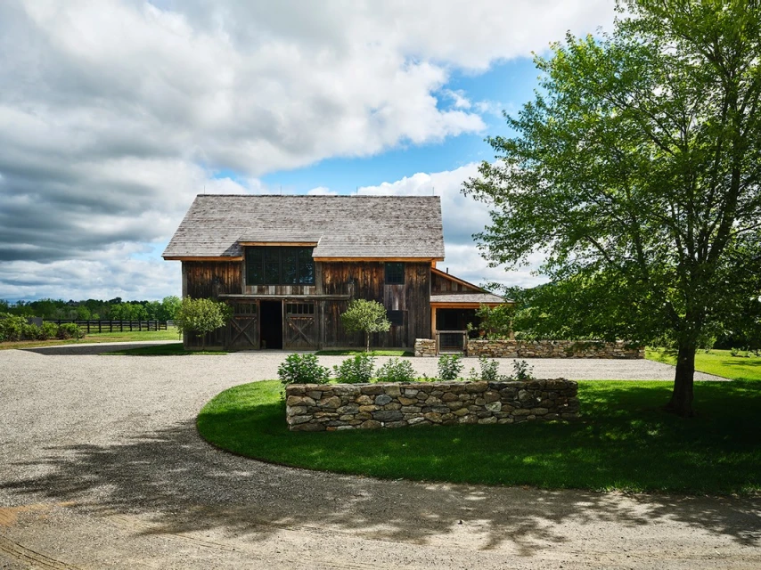 Rustic barn with weathered wood and grey roof surrounded by lush green grass and gravel driveway on cloudy day in rural setting.