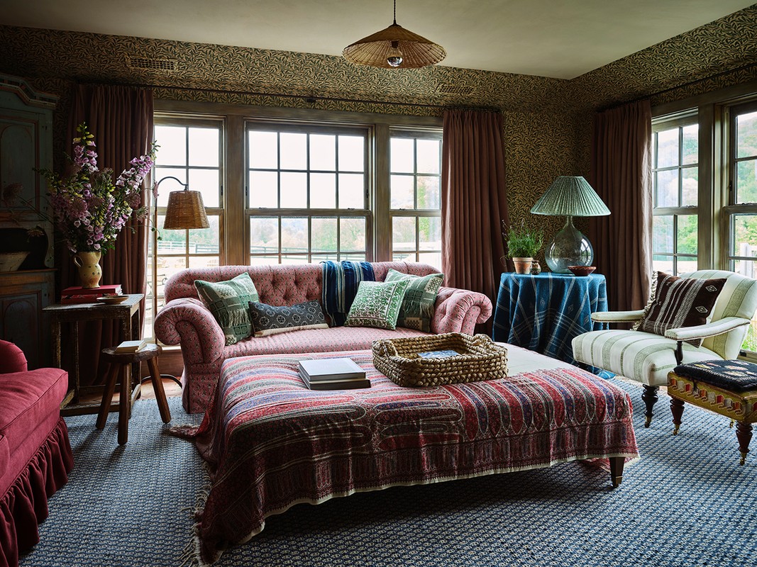 Cozy interior living room with patterned wallpaper, pink sofa, patterned ottoman, floral arrangement, table lamps, and multiple windows.