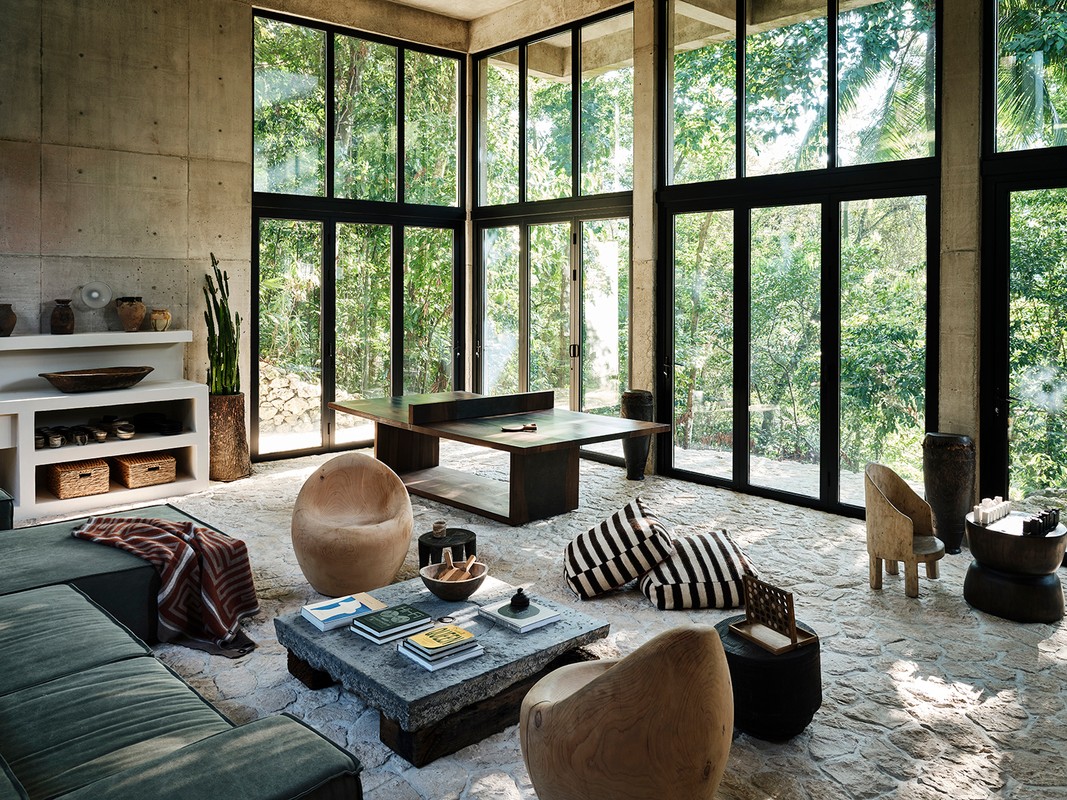 Spacious interior with concrete walls, floor-to-ceiling windows offering forest views, and modern furniture in a natural color palette.