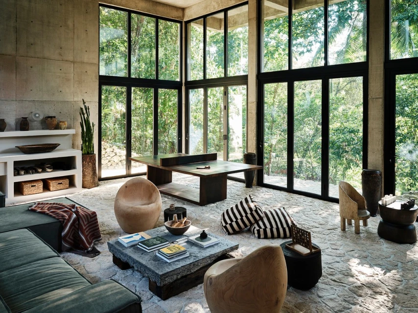 Spacious interior with concrete walls, floor-to-ceiling windows offering forest views, and modern furniture in a natural color palette.