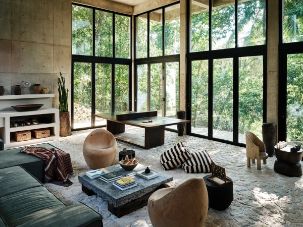 Spacious interior with concrete walls, floor-to-ceiling windows offering forest views, and modern furniture in a natural color palette.