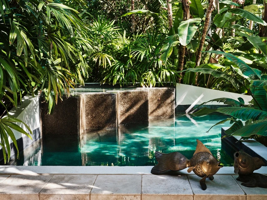 Lush tropical garden features a modern plunge pool with bronze fish sculptures and travertine tiles in natural daylight creating an oasis.