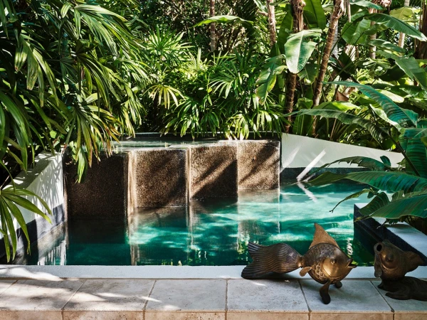 Lush tropical garden features a modern plunge pool with bronze fish sculptures and travertine tiles in natural daylight creating an oasis.