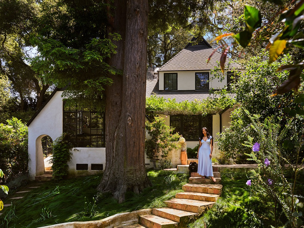Woman in a long dress stands on stone steps in front of a white house with a black roof, surrounded by trees and foliage in a yard.
