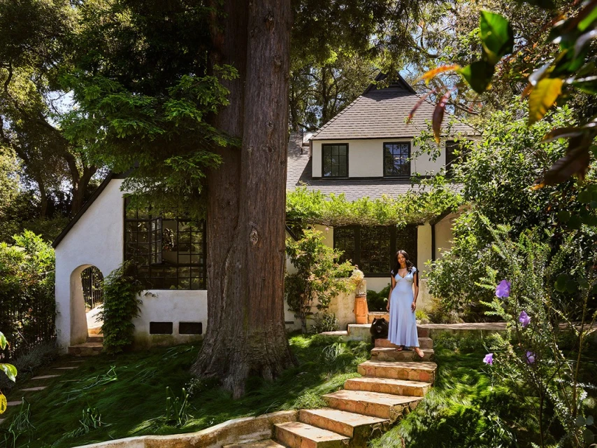 Woman in a long dress stands on stone steps in front of a white house with a black roof, surrounded by trees and foliage in a yard.