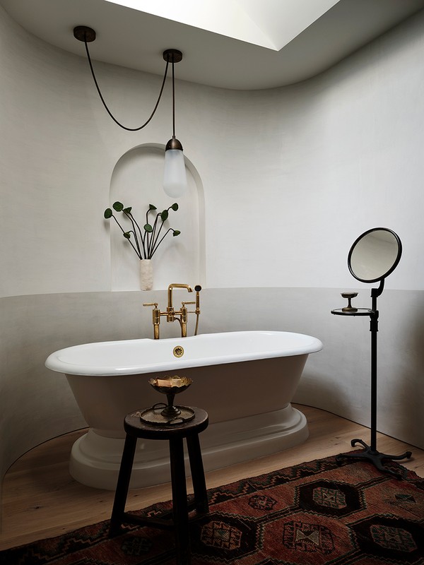 Bathroom interior with a freestanding white bathtub, black accent stool, round accent mirror, and a dark patterned rug on a light wood floor.