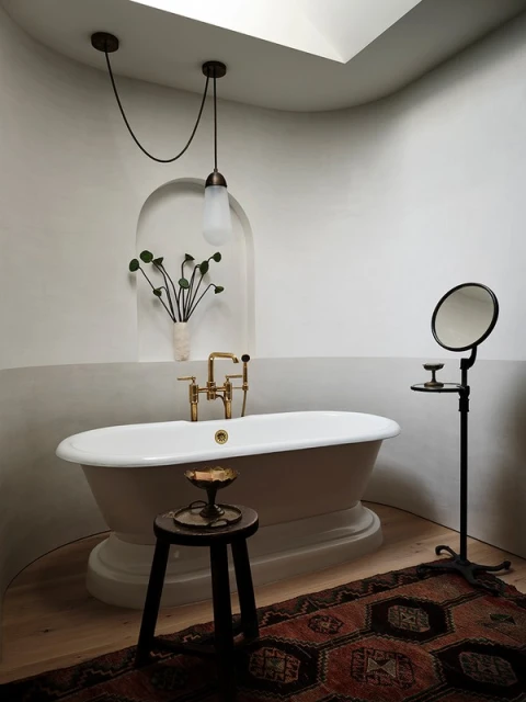 Bathroom interior with a freestanding white bathtub, black accent stool, round accent mirror, and a dark patterned rug on a light wood floor.