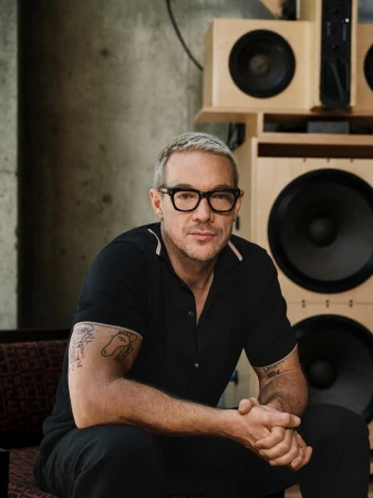 Man with short bleached hair wearing black polo and glasses, seated with hands clasped together, speaker boxes stacked behind him.