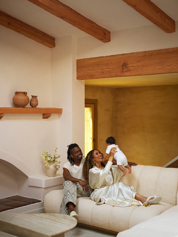 Family portrait in a living room: a man and woman are on a beige couch, holding a baby. Decorative vases and beams visible.