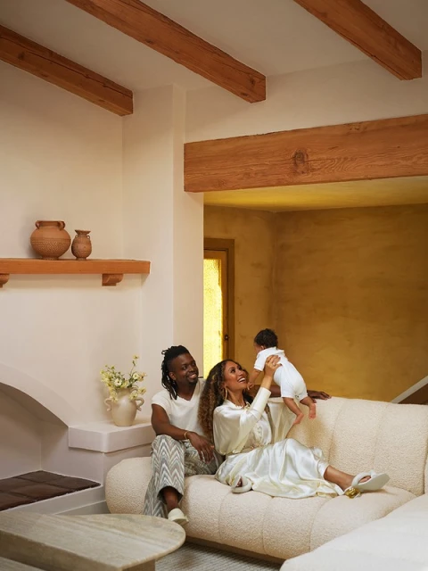 Family portrait in a living room: a man and woman are on a beige couch, holding a baby. Decorative vases and beams visible.