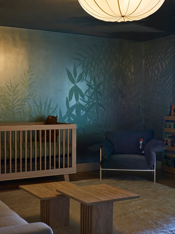 Nursery interior featuring a wooden crib, blue armchair, two wooden coffee tables, and a leafy green wall with shadowy accents.