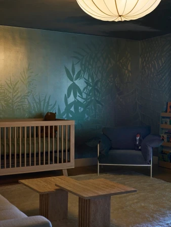 Nursery interior featuring a wooden crib, blue armchair, two wooden coffee tables, and a leafy green wall with shadowy accents.