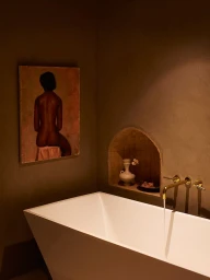 White freestanding bathtub with running brass faucet beneath an arched wall niche and a framed painting on a dark plaster wall.